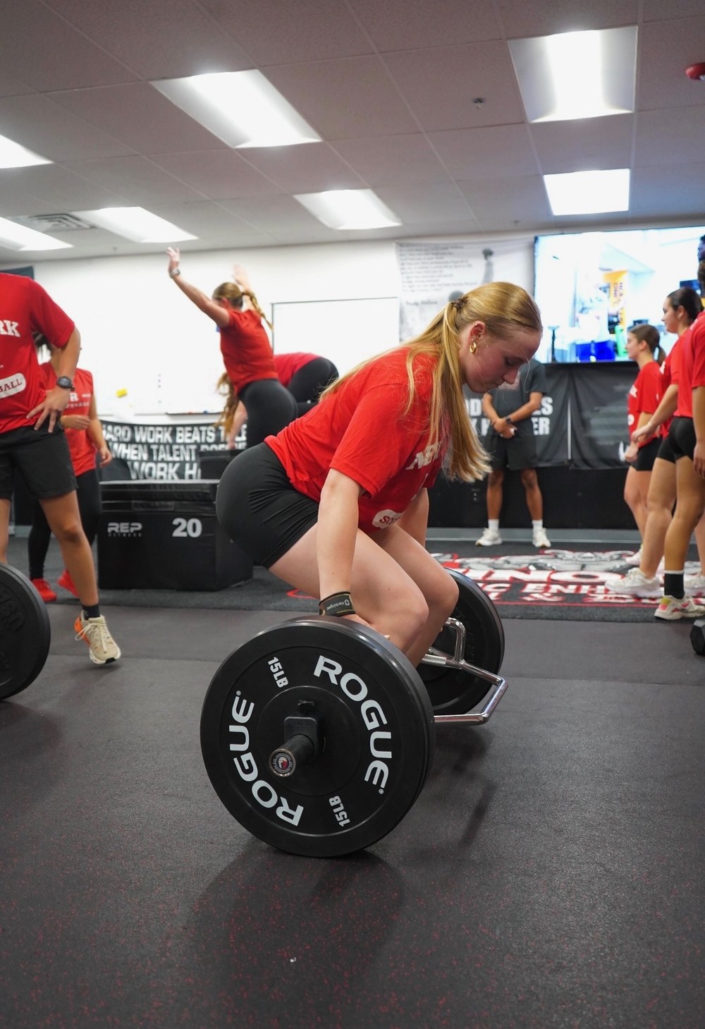 Athlete in a red shirt lifting a loaded Rogue barbell during a team session in the gym